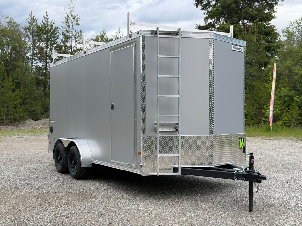 Silver enclosed cargo trailer with two axles, side door, and a rear ladder on the gravel lot, trees in the background.