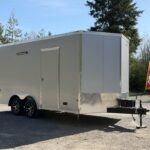 White enclosed cargo trailer with dual axles and a front hitch on a gravel lot.