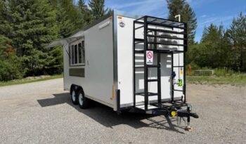 White utility trailer on a gravel lot with a metal rear ladder/rack, two-axle wheels, and trees in the background.