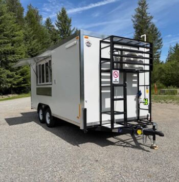 White utility trailer on a gravel lot with a metal rear ladder/rack, two-axle wheels, and trees in the background.