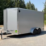 Enclosed silver cargo trailer with tandem axles on a gravel lot, hitch and jack at the front, Haulmark logo on top edge, trees in the background
