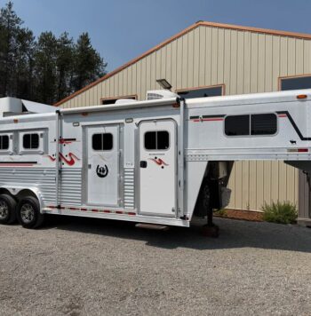 White gooseneck horse trailer with living quarters, four axles, and side windows parked on gravel near a beige metal building; red/black decals visible; horses transported inside.
