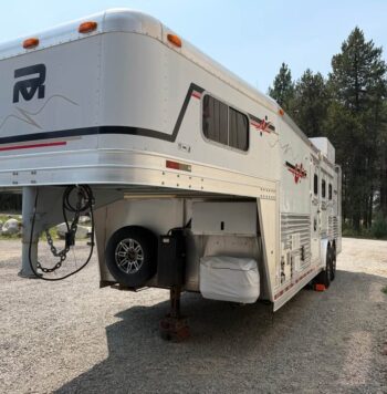Large white fifth-wheel travel trailer parked on gravel, its front raised on jacks with a spare tire and storage beneath. Nearby trees and a clear sky in the background.
