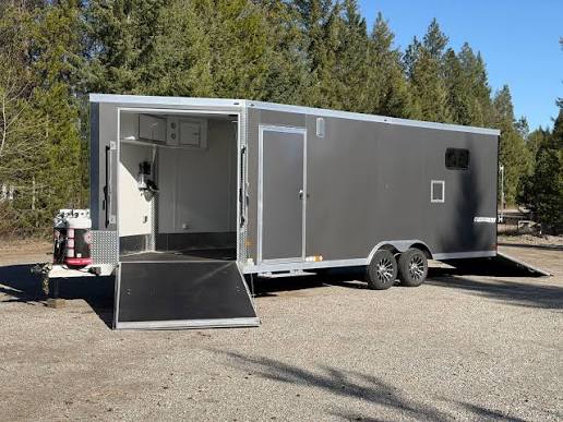 Silver utility trailer with open back ramp, showing interior cabinets and equipment, parked on gravel with trees in background.