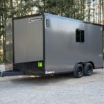 Gray Haulmark trailer with dual axles on a gravel lot, hitch at front and green number 16 on the front panel, forest behind.
